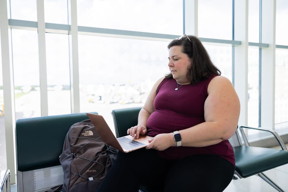 A woman sits in an airport with a backpack in front of her and a laptop partially propped up on her lap and on her backpack.