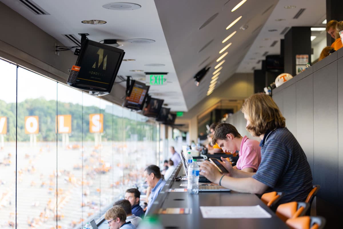 Students work on their laptops inside the press box at Neyland Stadium, the iconic Vols letters visible through the glass windows.