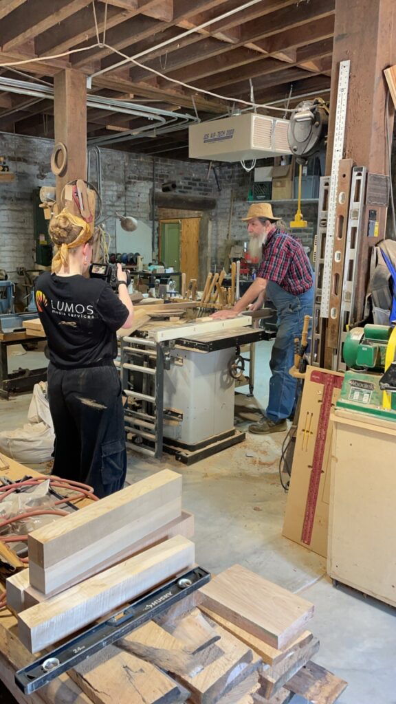 A student holds a camera and films a woodworker working on a project in his workshop.