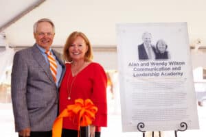 Alan Wilson, wearing a gray-brown suit coat and a UT orange and white tie with a blue button-up Oxford, stands with Wendy Wilson, wearing a red long-sleeve shirt, next to a plaque with their likenesses and a description of the Alan and Wendy Wilson Communication and Leadership Academy on it.