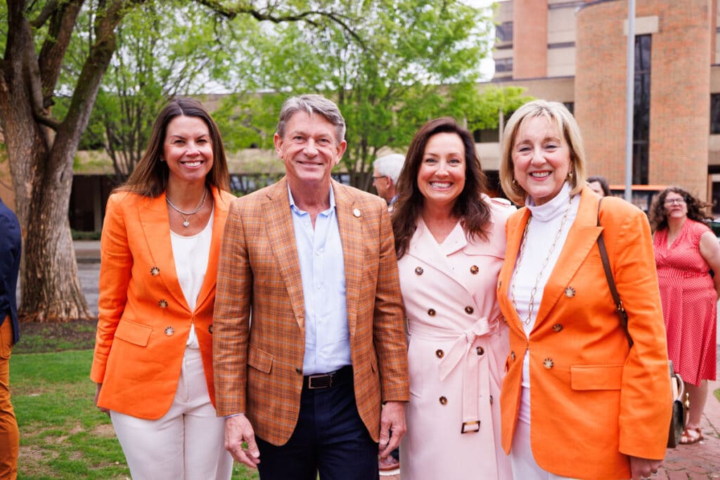 Two women wearing the exact same outfit of white shirt and pants and an orange sports caught stand on the right and left of two other people. On the middle left is a man wearing an orange plaid sports coat and on the middle right is a woman wearing a light pink long jacket.