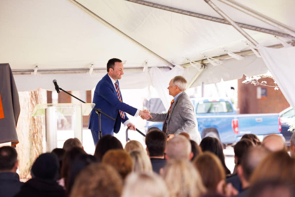 Joe Mazer, in a navy blue suit, is on a stage under a large white tent and bending over slightly to shake hands with Alan Wilson, who is coming up the stage's steps.