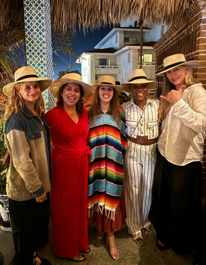 Five people pose beneath a grass roof structure, all of them wearing straw hats.