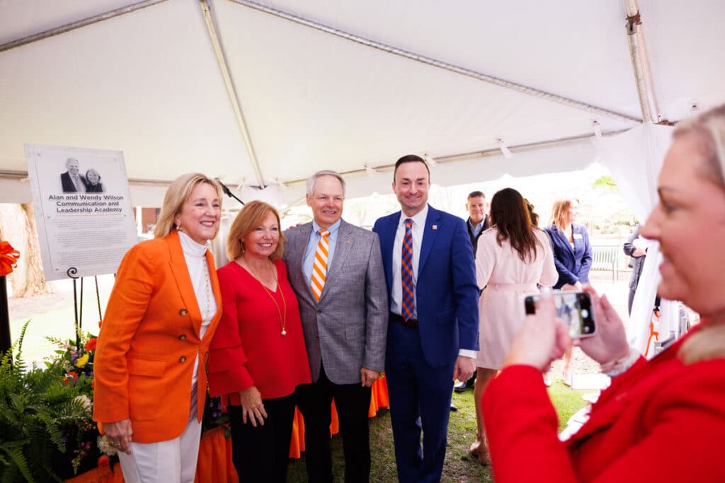Four people stand under a large white tent and post for a photo in front of a plaque with Alan and Wendy Wilson's likeness on it.