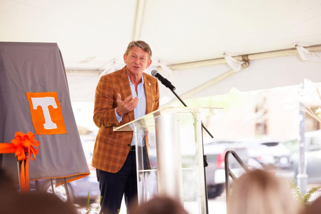 U-T System President Randy Boyd speaks and stands behind a clear podium by an easel with a grey blanket over it featuring a white power T on an orange square in the middle of it.