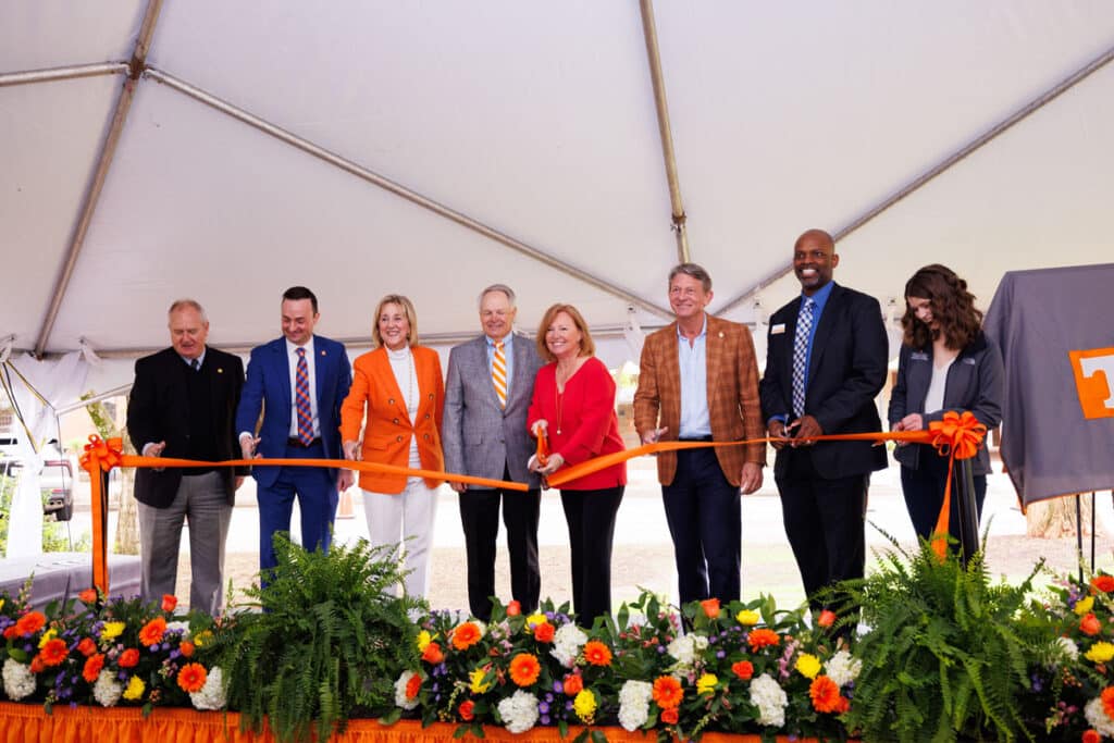 Eight people on a stage under a large white tent gather in front of a UT orange ribbon that Wendy Wilson is cutting with large scissors.