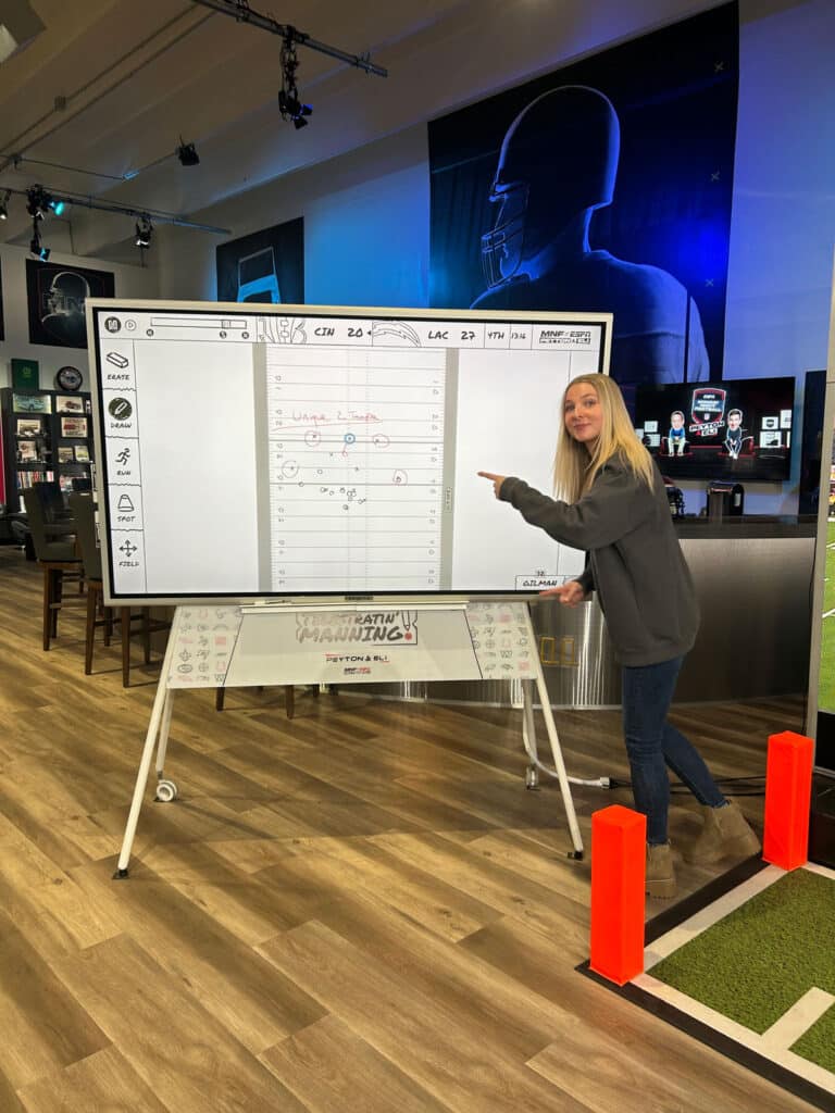 Addison Lafayette stands in front of a white board with plays written on it, pointing to it and smiling.