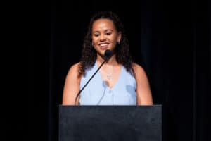 Riley West wears a light blue sleeveless button up shirt while standing behind a black podium with a microphone in front of her. The background is black.