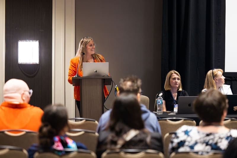 A woman wearing an orange sports coat stands behind a podium as she gives a presentation.