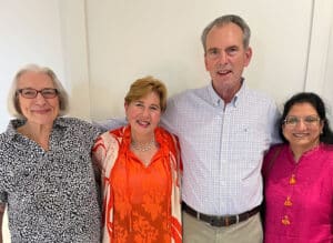 Four adults stand with their arms around each other in front of a white wall.