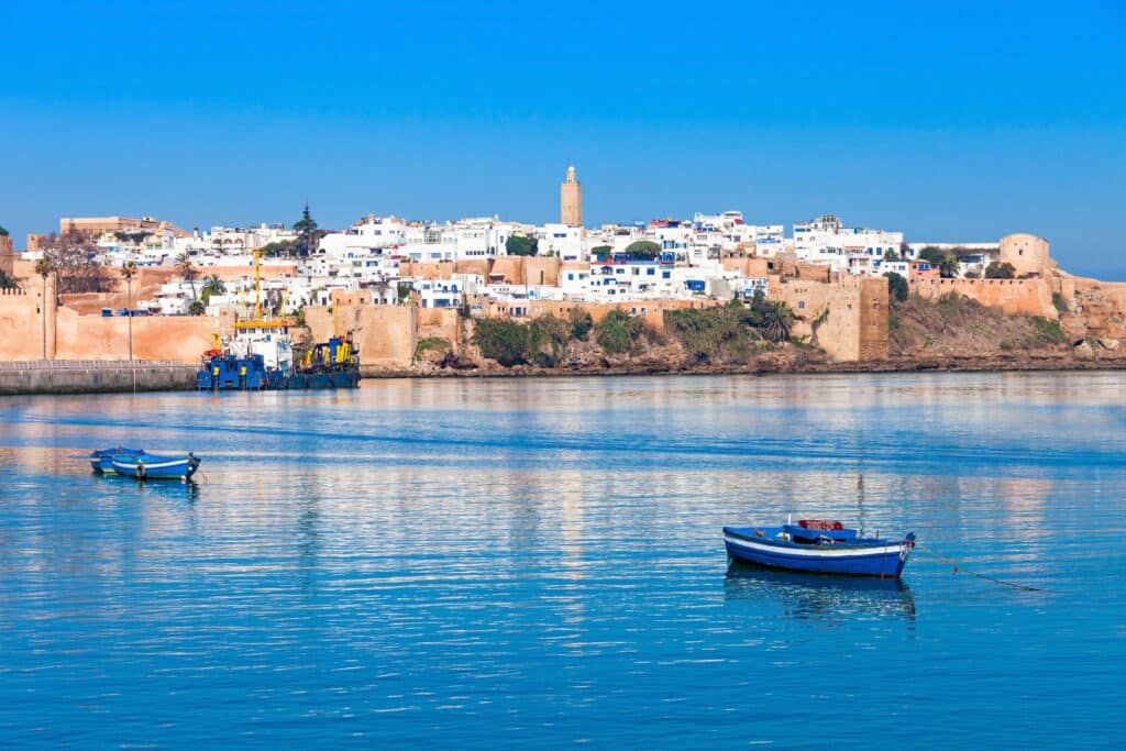 A city, Rabat, Morocco, comprised of white and tan buildings is set behind bright blue sea with fishing boats on it.