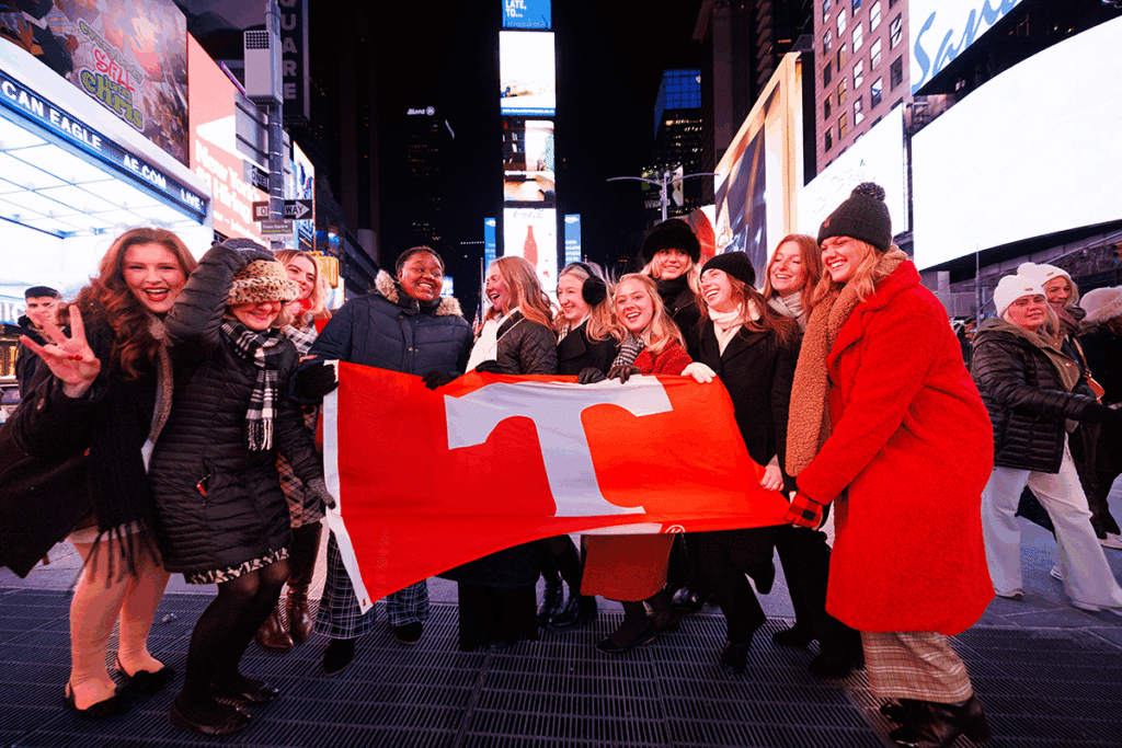 A picture of a group of School of Journalism and Media students posing with he Power T flag in Times Square at. night during the 2025 school trip to New York City.