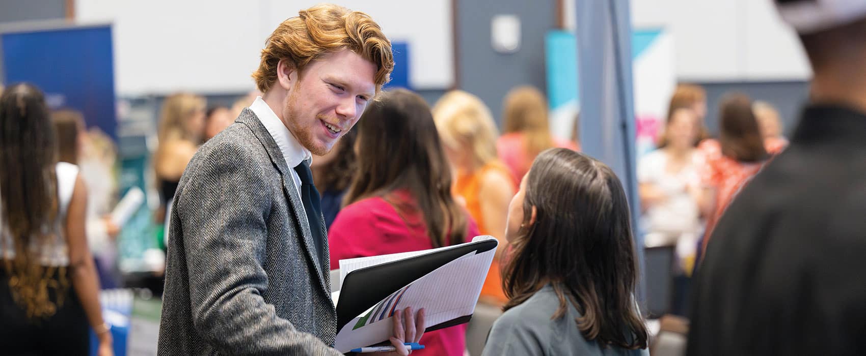 A student in a gray suit, white shirt, and tie smiles and shakes the hand of an employer in the middle of booths at a C-C-I career fair.