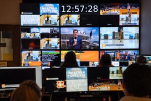 Silhouettes of students are seen as they sit in front of about 30 screens stacked together in a square in front of them in a T-V production room.