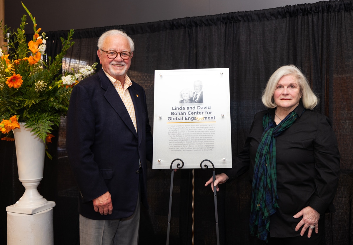 A plaque sits on a stand and features the story of the David and Linda Bohan Center for Global Engagement and David stands to the left of it and Linda to the right. There is a tall, white flower vase with orange and white flowers in it to the left of David.