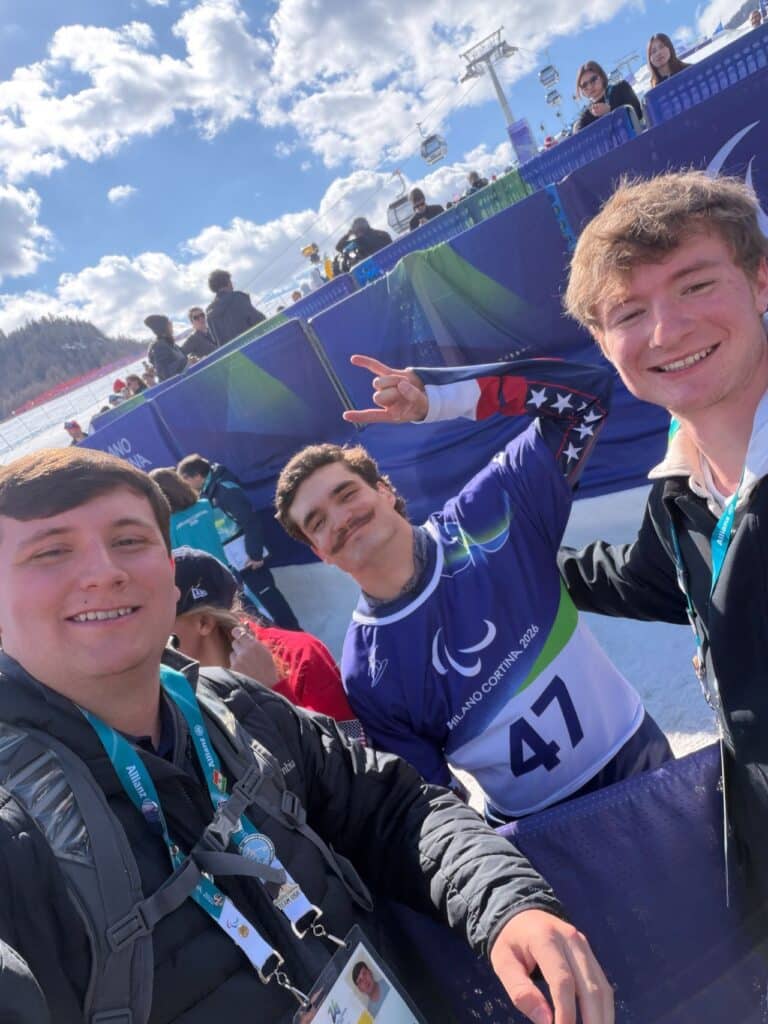 Selfie with a man on the left smiling wearing a black jacket and backpack with brown hair. The man in the middle is wearing a Milano Cortina jersey with his hands up holding two fingers with brown hair and a brown mustache. The man on the right has blonde hair a wearing a black jacket and smiling. They are at the winter games in Milan. There is snow in the background and a blue sky with clouds and some other people standing around in the background.