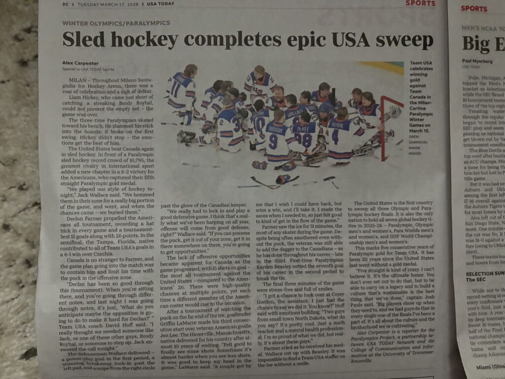 Photo of a newspaper clipping. The heading reads Sled hockey completes epic USA sweep. The byline reads Alex Carpenter. There is an image on the newspaper clipping of Team U-S-A celebrating a gold win at the Paralympics Game wearing U-S-A jerseys on the ice rink.