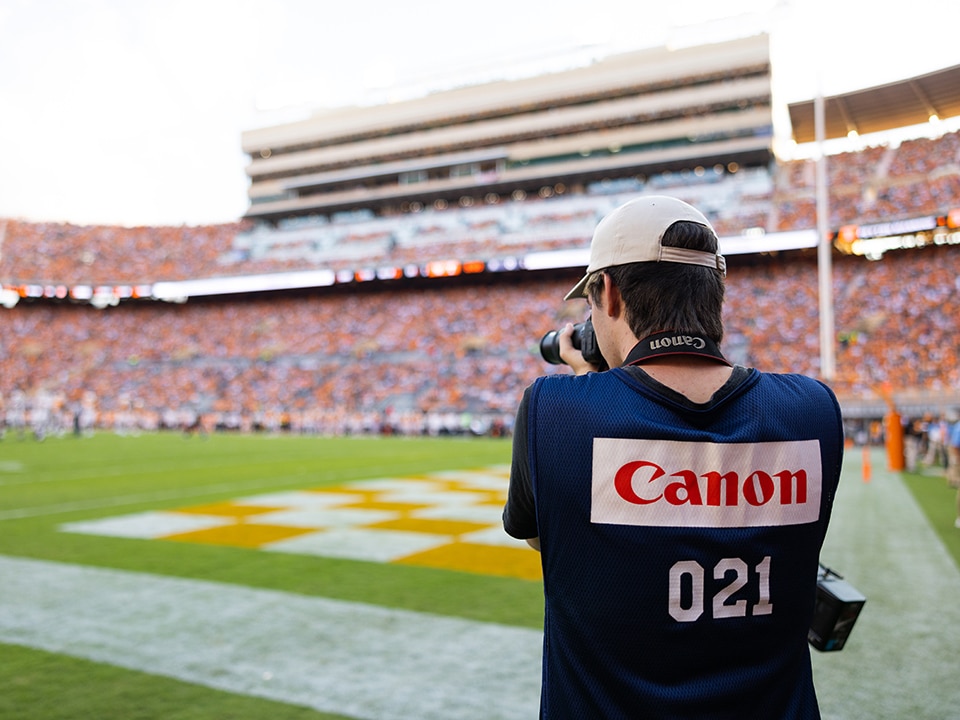 A student wearing a blue Canon shirt stands with his back to the viewer while he uses a camera to take photos of a football game at Neyland stadium, with the checkerboard endzone in view and orange clad fans in the seats.