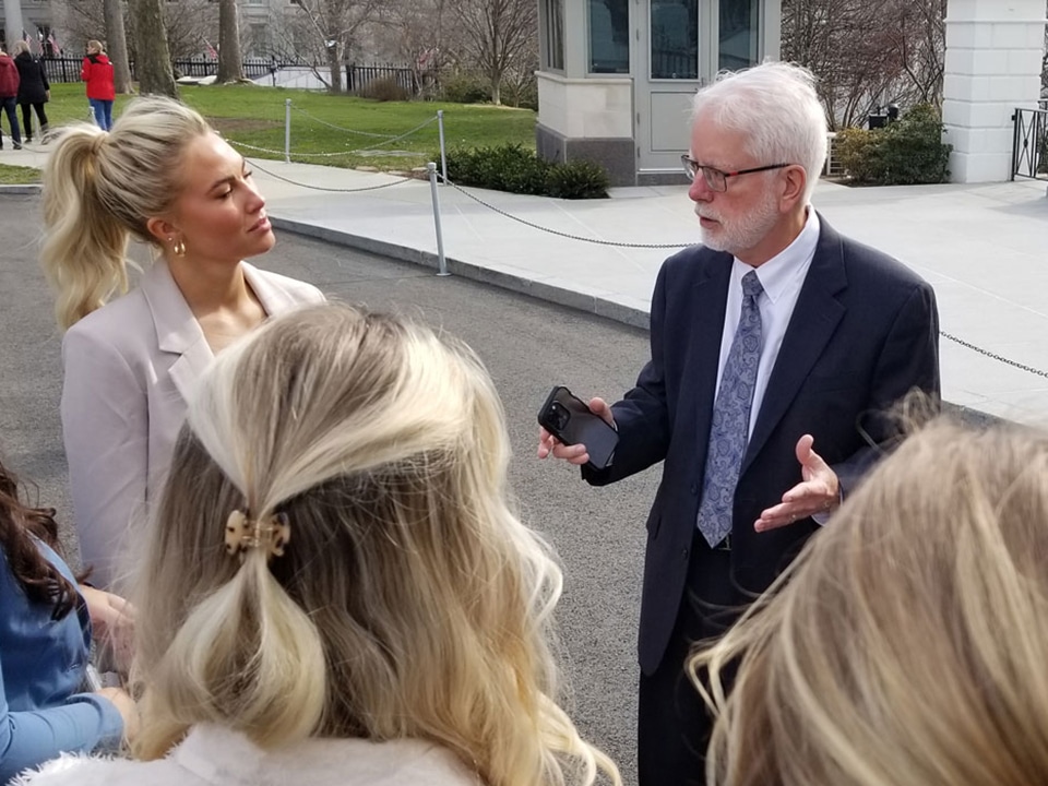 A professor wearing a suit and tie is surrounded by a handful of students as they stand in front of Washington, D-C sights.