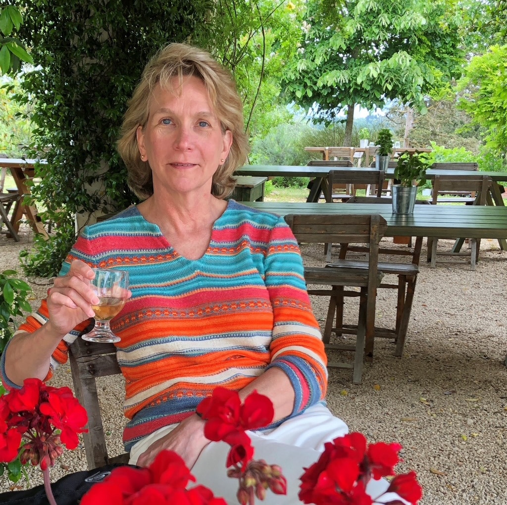 Graduating College of Communication and Information master's student Laura Ward in a colorful striped sweater sits outdoors, holding a glass, with red geraniums in the foreground and wooden tables in the background.