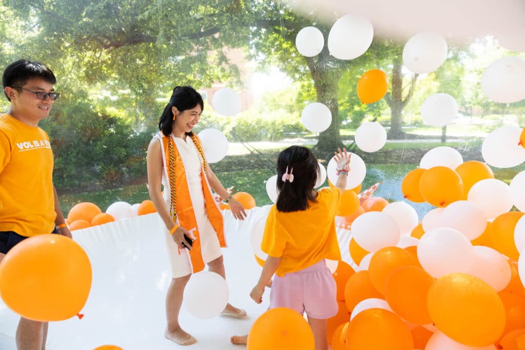 A graduate student plays with orange and white balloons with her daughter and spouse at the C-C-I send off.