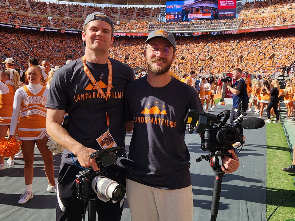 Two male students holding camera and video equipment stand on the turf in Neyland Stadium, which has a full crowd.