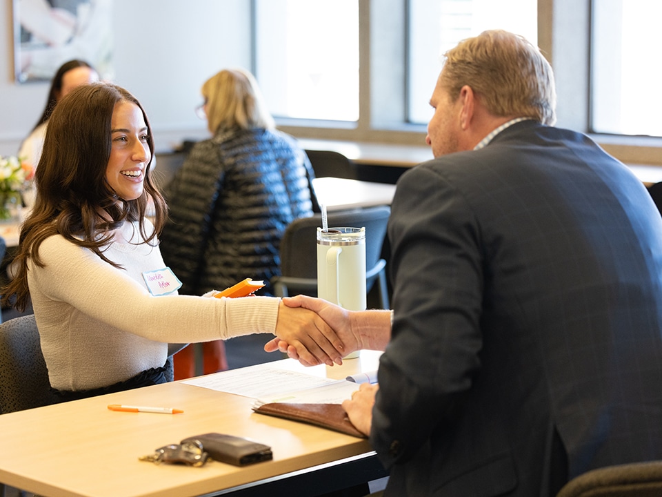 A student sits across the table from an employer and shakes his hand.